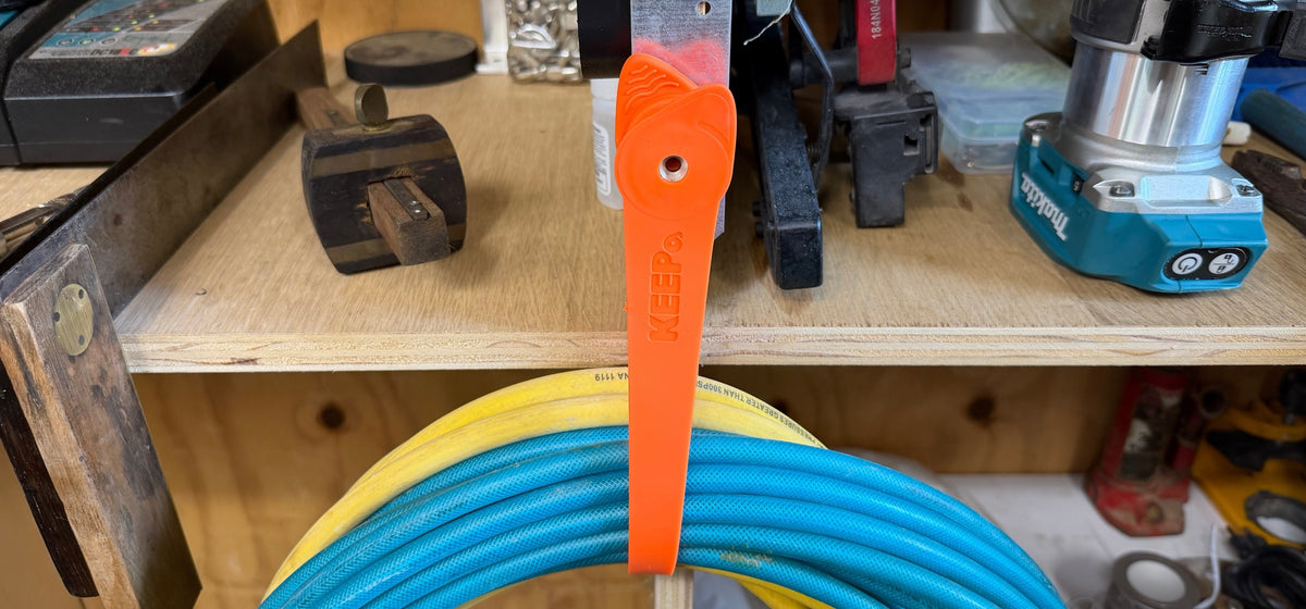 An orange KEEPa 14 clipped by its magnet to a tool on a workshop shelf, its strap holding a coiled hose.